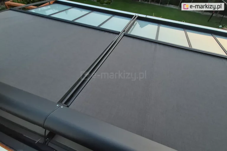 Zdjęcie przedstawia widok z góry na system dwumodułowej markizy werandowa, zamontowanej na aluminiowej konstrukcji ogrodu zimowego. Poszycie markizy wykonane jest z wysokiej jakości tkaniny akrylowej o gęstym splocie w kolorze ciemnoszarym (antracytowym). System jest w fazie częściowego rozwinięcia – dwa segmenty materiału są wysunięte do połowy, odsłaniając w górnej części kadru przeszklone panele dachu ujęte w czarne ramy. Elementy konstrukcyjne, w tym zaokrąglona kaseta osłonowa, prowadnice boczne oraz belki frontowe, są lakierowane proszkowo na kolor matowego antracytu. W tle widoczny jest fragment zadbanego ogrodu z zielonym trawnikiem, drzewami liściastymi oraz drewnianą konstrukcją pergoli po lewej stronie. Perspektywa zdjęcia z góry pozwala na dokładne przyjrzenie się mechanizmowi prowadzenia tkaniny oraz estetyce wykończenia detali.