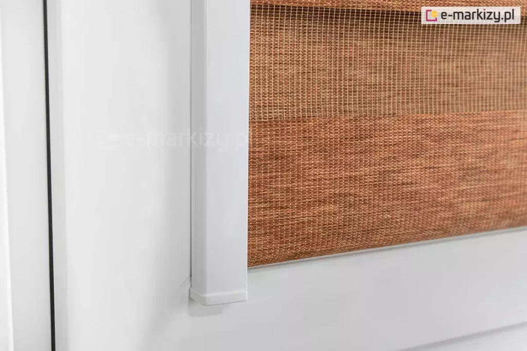 Zbliżenie na dolny narożnik systemu rolety lux kombi zamontowanej na białej ramie okiennej wykonanej z PVC. Zdjęcie przedstawia precyzyjne wykończenie pionowej prowadnicy bocznej, która jest wykonana z gładkiego aluminium w kolorze klasycznej bieli i zakończona dopasowaną estetyczną zaślepką z tworzywa sztucznego. Prowadnica przylega bezpośrednio do ramy okna, zapewniając stabilne prowadzenie materiału i eliminując prześwity boczne. Zastosowana w rolecie lux kombi tkanina dzień i noc składa się z poziomych pasów o zróżnicowanej przepuszczalności światła. Widoczny jest pas zaciemniający o melanżowej strukturze w ciepłym odcieniu miedzianego brązu, który charakteryzuje się gęstym splotem włókien o naturalnym wyglądzie. Powyżej znajduje się pas transparentny wykonany z delikatnej siatki o drobnych oczkach w podobnej tonacji kolorystycznej, która pozwala na wgląd na zewnątrz przy zachowaniu prywatności. Materiał rolety lux kombi chowa się pod krawędź bocznej prowadnicy, co gwarantuje estetyczny wygląd i funkcjonalność osłony nawet przy uchylonym skrzydle okiennym. Całość prezentuje nowoczesny design i wysoką jakość wykonania detali konstrukcyjnych systemu.