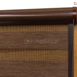 Zbliżenie na górną część wewnętrznej osłony okiennej typu roleta lux kombi zamontowanej na ramie okna o strukturze imitującej naturalne drewno w jasnym odcieniu dębu. Produkt charakteryzuje się naprzemiennym układem poziomych pasów wykonanych z dwóch różnych materiałów, co pozwala na precyzyjną regulację dopływu światła do pomieszczenia. Pasy pełne mają brązowy kolor o wyraźnej, tekstylnej strukturze z delikatnym prążkowaniem, natomiast pasy transparentne wykonane są z drobnej, gęstej siatki w kolorze miodowym lub złocistym, która przepuszcza rozproszone światło. Cały mechanizm rolety lux kombi ukryty jest w eleganckiej, zaokrąglonej kasecie osłonowej wykonanej z gładkiego aluminium w kolorze ciemnego brązu o satynowym wykończeniu. Kaseta jest zamontowana bezpośrednio przy górnej krawędzi skrzydła okiennego i zakończona dopasowanym kolorystycznie boczkiem z tworzywa. Z prawej strony boczka wychodzi łańcuszek kulkowy służący do sterowania wysokością rolety oraz wzajemnym położeniem pasów tkaniny dzień noc względem siebie. Po bokach tkaniny widoczne są pionowe prowadnice w kolorze ciemnobrązowym, które utrzymują materiał rolety lux kombi blisko szyby, zapobiegając jego odchylaniu się przy uchylonym oknie i zapewniając płynną pracę mechanizmu.