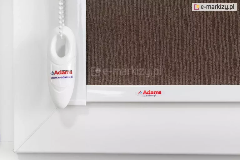 Zbliżenie lewego narożnika skrzydła okna o białej ramie z widoczną prowadnicą przestrzenną, obciążnikiem dolnym aluminiowym oraz łańcuszkiem z obciążnikiem, na którym umieszczono naklejkę producenta. Widoczna także tkanina rolety z lekko strukturalnym wzorem w nieregularne pionowe linie w kolorze brązowym.