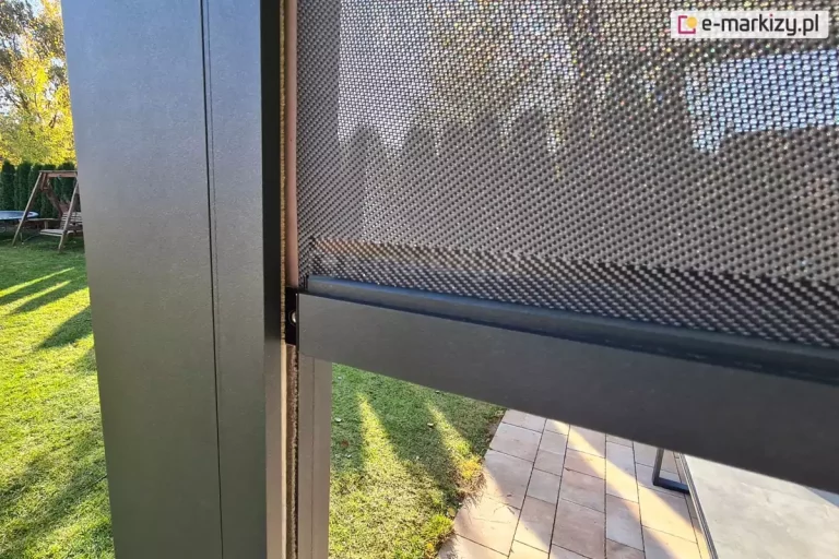 Zbliżenie na boczny profil prowadnicy oraz dolną listwę obciążającą zewnętrznego refleksola Zip z tkaniną typu screen zainstalowanej przy nowoczesnej konstrukcji tarasowej. Na zdjęciu widoczna jest pionowa, aluminiowa noga pergoli w kolorze ciemnego antracytu o matowym wykończeniu, do której przymocowana jest prowadnica boczna zintegrowana z systemem zamkowym tkaniny. Przezroczysta, techniczna tkanina o drobnym splotu siatkowym w odcieniach szarości i czerni jest częściowo rozwinięta, ukazując strukturę mikrootworów zapewniających ochronę przeciwsłoneczną przy jednoczesnym zachowaniu widoczności na zewnątrz. Dolna krawędź materiału jest estetycznie zakończona poziomą, masywną listwą obciążającą wykonaną z aluminium, malowaną proszkowo na ten sam odcień co konstrukcja. Listwa ta jest precyzyjnie osadzona w bocznych prowadnicach, co gwarantuje stabilność materiału nawet przy podmuchach wiatru. W tle zdjęcia znajduje się nasłoneczniony ogród z zielonym trawnikiem, fragmentem drewnianej huśtawki ogrodowej oraz ułożoną kostką brukową na tarasie, na którą padają wyraźne cienie rzucane przez konstrukcję w niskim, popołudniowym świetle.