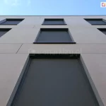 Rolety ZIP L zamocowane w oknach trzech pięter budynku o szarej betonowej fasadzie. Rolety z antracytową tkaniną są pozamykane, tworząc wewnątrz przyjemny cień.