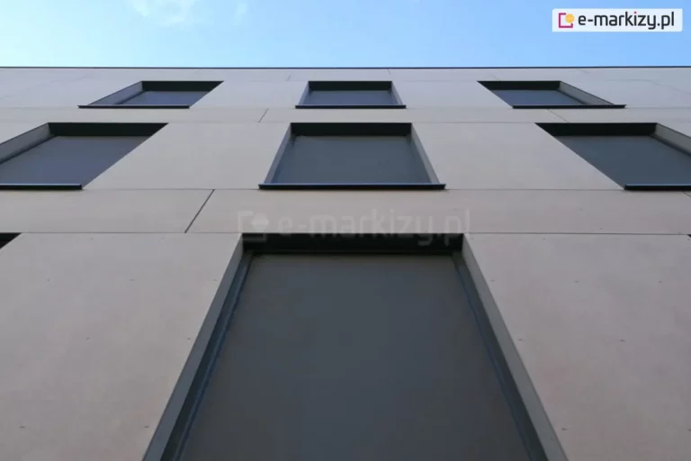 Rolety ZIP L zamocowane w oknach trzech pięter budynku o szarej betonowej fasadzie. Rolety z antracytową tkaniną są pozamykane, tworząc wewnątrz przyjemny cień.