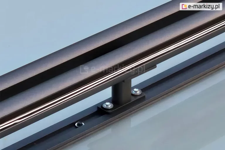 Uchwyt modułowy systemu veranda, który pozwala zamocować obok siebie dwie prowadnice rolety. Uchwyt przykręcony do krokwi dachowej zadaszenia w kolorze antracytowym. Ponad uchwytem widoczna wewnętrzna część prowadnicy z zamocowaną w niej białą linką.