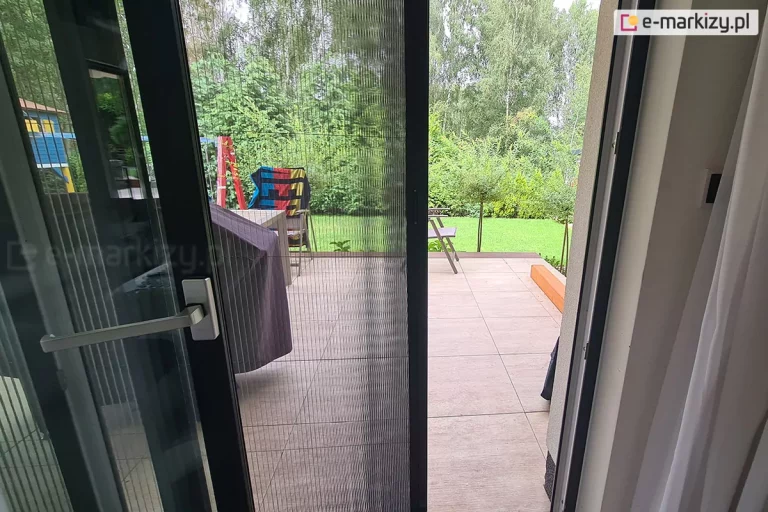 Uchylone drzwi tarasowe o przesuwnym skrzydle w kolorze czarnym ze srebrną klamką osłonięte częściowo moskitierą plisowaną. Na tarasie za oknem widoczne meble tarasowe ustawione na płytkach gresowych, częściowo przesłonięte zawieszonymi na nich materiałami. W tle zielony trawnik i krzewy.