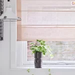 Zbliżenie na fragment jasnej kuchni, w której centralnym punktem jest okno przesłonięte przez delikatne rolety rzymskie. Tkanina osłony ma subtelny, pastelowy odcień brzoskwiniowy lub pudrowy róż i charakteryzuje się lekką, półtransparentną strukturą z wyraźnie widocznym splotem przypominającym naturalny len. Rolety rzymskie są częściowo podciągnięte, a poziome fiszbiny dzielą materiał na estetyczne, równoległe sekcje, które u dołu tworzą miękką zakładkę. Przez transparentny materiał do wnętrza wpada rozproszone światło dzienne, a w tle majaczy zarys ceglanego muru znajdującego się na zewnątrz. Okno posiada białą ramę i osadzone jest w gładkiej, białej ścianie. Na białym parapecie, bezpośrednio pod roletą, ustawiono dekoracyjną, ciemnoszarą doniczkę o rzeźbiarskiej formie przypominającej twarz, z której wyrasta zielona roślina o drobnych, pnących liściach. Po lewej stronie zdjęcia widoczny jest fragment białej lampy o industrialnym kształcie z metalowymi zatrzaskami oraz czarny, druciany regał ścienny, na którym umieszczono ceramiczne naczynia – w tym różowy dzbanek korespondujący kolorystycznie z tkaniną, z której wykonano rolety rzymskie. Całość kompozycji jest minimalistyczna, jasna i utrzymana w spokojnej, nowoczesnej estetyce.