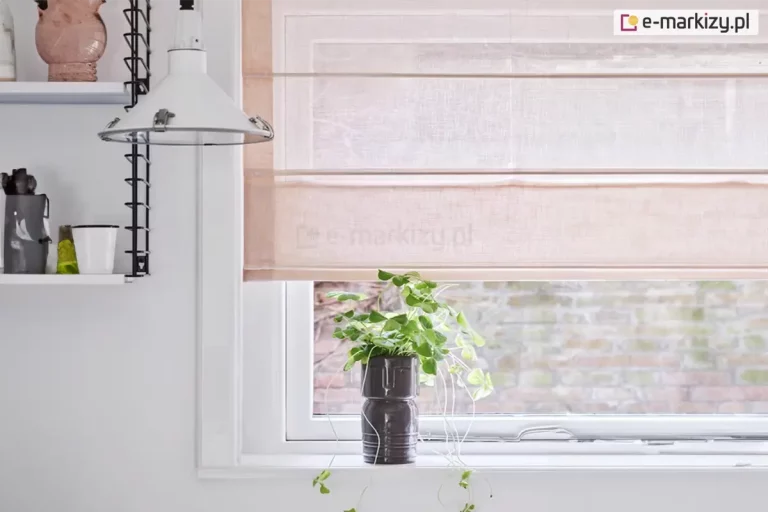 Zbliżenie na fragment jasnej kuchni, w której centralnym punktem jest okno przesłonięte przez delikatne rolety rzymskie. Tkanina osłony ma subtelny, pastelowy odcień brzoskwiniowy lub pudrowy róż i charakteryzuje się lekką, półtransparentną strukturą z wyraźnie widocznym splotem przypominającym naturalny len. Rolety rzymskie są częściowo podciągnięte, a poziome fiszbiny dzielą materiał na estetyczne, równoległe sekcje, które u dołu tworzą miękką zakładkę. Przez transparentny materiał do wnętrza wpada rozproszone światło dzienne, a w tle majaczy zarys ceglanego muru znajdującego się na zewnątrz. Okno posiada białą ramę i osadzone jest w gładkiej, białej ścianie. Na białym parapecie, bezpośrednio pod roletą, ustawiono dekoracyjną, ciemnoszarą doniczkę o rzeźbiarskiej formie przypominającej twarz, z której wyrasta zielona roślina o drobnych, pnących liściach. Po lewej stronie zdjęcia widoczny jest fragment białej lampy o industrialnym kształcie z metalowymi zatrzaskami oraz czarny, druciany regał ścienny, na którym umieszczono ceramiczne naczynia – w tym różowy dzbanek korespondujący kolorystycznie z tkaniną, z której wykonano rolety rzymskie. Całość kompozycji jest minimalistyczna, jasna i utrzymana w spokojnej, nowoczesnej estetyce.