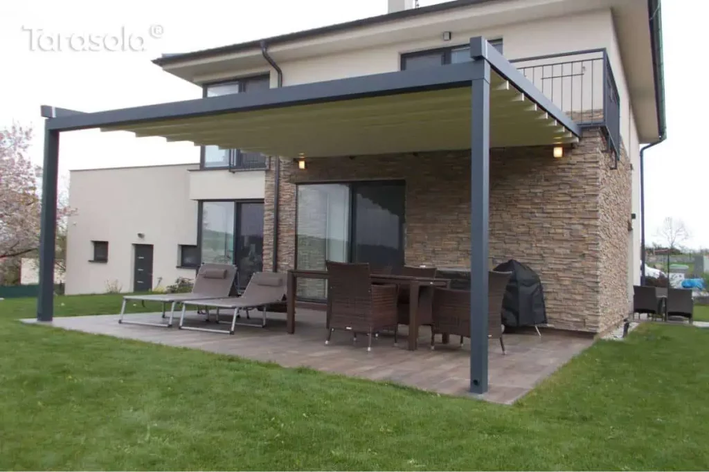 Przyścienna pergola tarasowa Elegancy Tarasola z całkowicie rozwiniętym dachem rzymskim, zamontowana przy nowoczesnym budynku z elewacją wykończoną dekoracyjnym kamieniem. Konstrukcja aluminiowa w kolorze antracytowym wspiera jasną tkaninę zadaszenia, która chroni strefę wypoczynkową przed słońcem. Na tarasie wyłożonym płytkami o strukturze drewna znajduje się zestaw mebli ogrodowych z ciemnego technorattanu oraz dwa szare leżaki z zagłówkami. W rogu ustawiono grill pod czarnym pokrowcem, a nad oknem tarasowym widoczne są zamontowane punkty oświetlenia zewnętrznego. Całość otacza zadbany zielony trawnik, a w oddali widoczne są kwitnące drzewa owocowe oraz pagórkowata okolica pod zachmurzonym niebem.