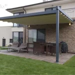 Przyścienna pergola tarasowa Elegancy Tarasola z całkowicie rozwiniętym dachem rzymskim, zamontowana przy nowoczesnym budynku z elewacją wykończoną dekoracyjnym kamieniem. Konstrukcja aluminiowa w kolorze antracytowym wspiera jasną tkaninę zadaszenia, która chroni strefę wypoczynkową przed słońcem. Na tarasie wyłożonym płytkami o strukturze drewna znajduje się zestaw mebli ogrodowych z ciemnego technorattanu oraz dwa szare leżaki z zagłówkami. W rogu ustawiono grill pod czarnym pokrowcem, a nad oknem tarasowym widoczne są zamontowane punkty oświetlenia zewnętrznego. Całość otacza zadbany zielony trawnik, a w oddali widoczne są kwitnące drzewa owocowe oraz pagórkowata okolica pod zachmurzonym niebem.