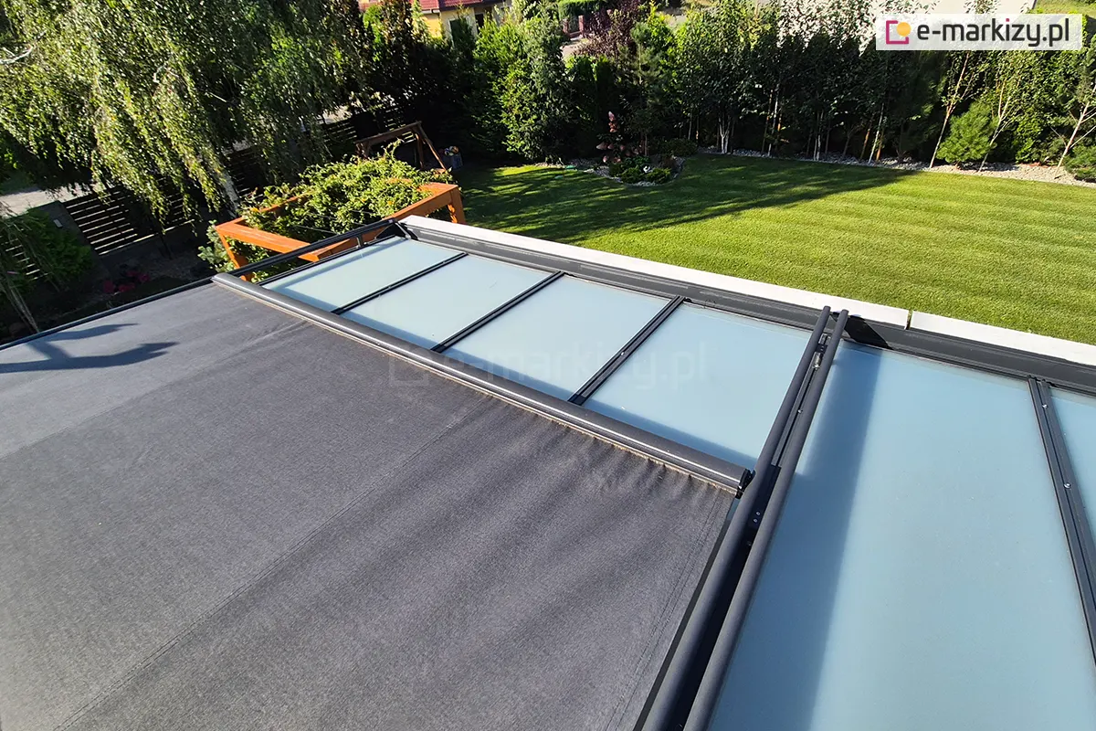 Zdjęcie przedstawia widok z góry na system veranda dwumodułowy, zamontowanych na dachu ogrodu zimowego. Poszycie markizy wykonane jest z wysokiej jakości tkaniny akrylowej w kolorze ciemnoszarym (antracytowym) o matowym wykończeniu. System jest modułowy – jeden segment verandy jest całkowicie rozwinięty, zasłaniając przeszklony dach, natomiast sąsiedni segment pozostaje odsłonięty, ukazując konstrukcję szklanych paneli ujętych w ciemne ramy. Aluminiowe prowadnice oraz profile systemu są lakierowane proszkowo na kolor antracyt. W tle rozpościera się widok na zadbany, zielony ogród z równo przyciętym trawnikiem, gęstą ścianą drzew iglastych oraz krzewami ozdobnymi. Widoczny jest również fragment drewnianej konstrukcji ogrodowej po lewej stronie. Całość ukazuje funkcjonalne zastosowanie markiz w ochronie dużych przeszkleń przed nagrzewaniem.