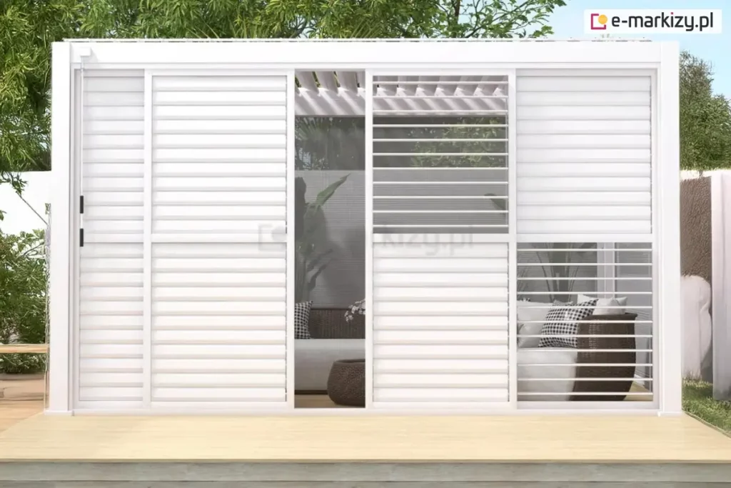 Zdjęcie prezentuje białą, aluminiową konstrukcję pergoli Gloria wyposażoną w system paneli żaluzjowych. Ściana boczna składa się z czterech paneli, z których dwa środkowe są rozsunięte, tworząc wejście do wnętrza strefy wypoczynkowej. Panele wypełnione są białymi, poziomymi lamelami, które w niektórych sekcjach są całkowicie domknięte, a w innych ustawione pod kątem, co pozwala na częściowy wgląd do środka i swobodną cyrkulację powietrza. Przez otwory widać wnętrze pergoli z meblami ogrodowymi, w tym sofą z poduszkami w kratkę i plecionym fotelem. Konstrukcja posiada płaski dach z widocznym systemem ruchomych lameli dachowych. Całość osadzona jest na jasnym tarasie wykonanym z desek kompozytowych, w otoczeniu ogrodowej zieleni i drzew liściastych widocznych w tle.