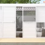 Zdjęcie prezentuje białą, aluminiową konstrukcję pergoli Gloria wyposażoną w system paneli żaluzjowych. Ściana boczna składa się z czterech paneli, z których dwa środkowe są rozsunięte, tworząc wejście do wnętrza strefy wypoczynkowej. Panele wypełnione są białymi, poziomymi lamelami, które w niektórych sekcjach są całkowicie domknięte, a w innych ustawione pod kątem, co pozwala na częściowy wgląd do środka i swobodną cyrkulację powietrza. Przez otwory widać wnętrze pergoli z meblami ogrodowymi, w tym sofą z poduszkami w kratkę i plecionym fotelem. Konstrukcja posiada płaski dach z widocznym systemem ruchomych lameli dachowych. Całość osadzona jest na jasnym tarasie wykonanym z desek kompozytowych, w otoczeniu ogrodowej zieleni i drzew liściastych widocznych w tle.