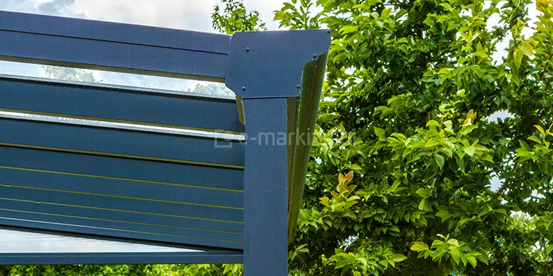 Detal konstrukcyjny aluminiowego zadaszenia tarasowego, skupiający się na systemie odprowadzania wody opadowej o klasycznym wykończeniu. Widoczne jest połączenie poziomej rynny frontowej z pionowym słupem nośnym o kwadratowym przekroju, wykonane z profili w kolorze antracytowym. Narożnik konstrukcji został zabezpieczony estetyczną maskownicą przymocowaną za pomocą wkrętów, która spaja rynnę ze słupem, zapewniając szczelność i sztywność układu. Nad rynną widoczne są zakończenia krokwi dachowych oraz zamontowane w nich przezroczyste panele szklane, które tworzą pokrycie dachu. Słup nośny pełni jednocześnie funkcję rury spustowej, ukrywając system drenażu wewnątrz profilu. W tle widoczne jest gęste, zielone ulistnienie drzewa oraz fragment jasnego nieba, co podkreśla ogrodowy charakter instalacji.