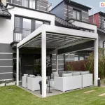 Nowoczesna pergola wolnostojąca z automatycznym dachem lamelowym, zamontowana przy tarasie domu szeregowego o minimalistycznej elewacji w biało-grafitowej kolorystyce. Konstrukcja pergoli wykonana z aluminiowych profili w kolorze białym, z dachem z ruchomych lameli ustawionych w pozycji otwartej. Pod pergolą znajduje się strefa wypoczynkowa z kompletem jasnych mebli ogrodowych z technorattanu – narożnikiem, fotelami i niskim stolikiem. Taras wyłożony płytami ceramicznymi, otoczony niskimi rabatami z roślinami ozdobnymi. W tle widać ogrodzenie z maty bambusowej, a całość zaaranżowana jest na zadbanym, zielonym trawniku przy nowoczesnym budynku wielorodzinnym z balkonami i grafitowymi dachami.