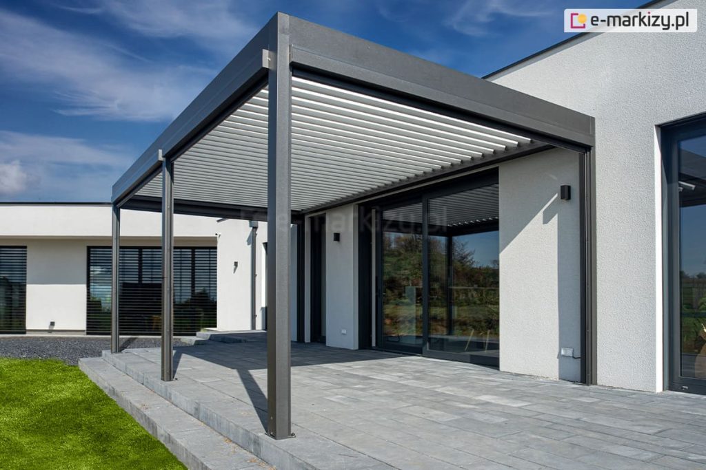 Bioklimatyczna pergola z otwartymi roletami i dachem ustawiona na tarasie betonowym przy parterowym domku o jasnej elewacji i grafitowej stolarce okiennej