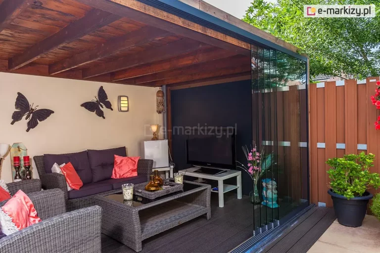 Nowoczesna aranżacja przydomowej strefy relaksu z wykorzystaniem bezramowego systemu szklanego Auris. Zdjęcie przedstawia narożną zabudowę drewnianej altany, w której frontowej ścianie zamontowano tafle szkła hartowanego przesuwane kaskadowo na wielotorowej prowadnicy dolnej. Wnętrze altany o ciemnym, drewnianym stropie wyposażone jest w luksusowe meble z szarego technorattanu (sofa, fotele, stolik kawowy ze szklanym blatem) oraz strefę multimedialną z telewizorem. Ściany zdobią dekoracyjne metalowe motyle, a nastrojowe oświetlenie zapewniają kinkiety. Zabudowa Auris skutecznie oddziela przestrzeń wypoczynkową od ogrodu, zapewniając ochronę przed wiatrem przy jednoczesnym zachowaniu pełnego wglądu na roślinność i brązowe ogrodzenie lamelowe.