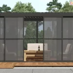 Nowoczesna, wolnostojąca pergola o konstrukcji aluminiowej Gloria, w kolorze antracytowym, wyposażoną w system przesuwnych. Ściany boczne wykonane są z dymionego poliwęglanu, ujętego w ciemne ramy z subtelnymi szprosami, co nadaje całości elegancki, industrialny wygląd. Jedno z przesuwnych skrzydeł drzwiowych jest otwarte, ukazując wnętrze strefy wypoczynkowej, w której znajduje się drewniany stolik kawowy oraz wygodna sofa z jasnymi poduszkami. Pergola została posadowiona na tarasie wykonanym z naturalnych desek o miodowym odcieniu, który kontrastuje z szarymi płytami chodnikowymi widocznymi na pierwszym planie. W tle rozpościera się widok na gęsty, słoneczny las iglasty pod bezchmurnym niebem. Konstrukcja dachu pergoli jest płaska, z widocznym od spodu systemem zadaszenia lamelowego o uchylonych piórach.