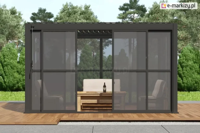 Nowoczesna, wolnostojąca pergola o konstrukcji aluminiowej Gloria, w kolorze antracytowym, wyposażoną w system przesuwnych. Ściany boczne wykonane są z dymionego poliwęglanu, ujętego w ciemne ramy z subtelnymi szprosami, co nadaje całości elegancki, industrialny wygląd. Jedno z przesuwnych skrzydeł drzwiowych jest otwarte, ukazując wnętrze strefy wypoczynkowej, w której znajduje się drewniany stolik kawowy oraz wygodna sofa z jasnymi poduszkami. Pergola została posadowiona na tarasie wykonanym z naturalnych desek o miodowym odcieniu, który kontrastuje z szarymi płytami chodnikowymi widocznymi na pierwszym planie. W tle rozpościera się widok na gęsty, słoneczny las iglasty pod bezchmurnym niebem. Konstrukcja dachu pergoli jest płaska, z widocznym od spodu systemem zadaszenia lamelowego o uchylonych piórach.