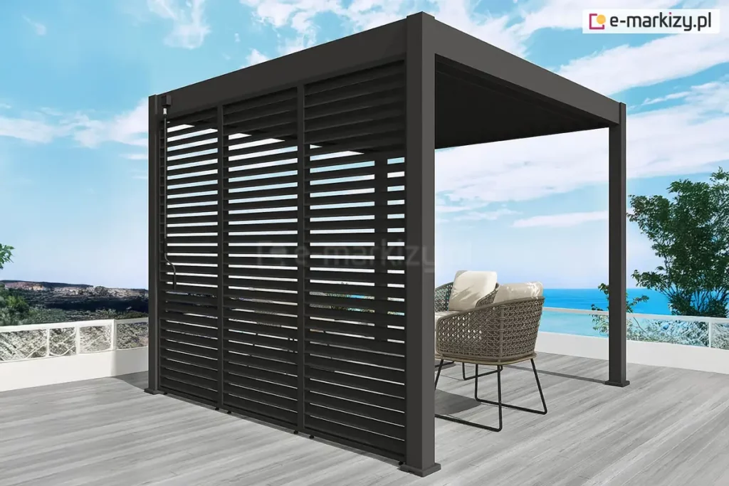 Antracytowa wolnostojąca pergola lamelowa Marlo o kwadratowej podstawie, usytuowana na tarasie z widokiem na morze. Konstrukcja wyposażona jest w pełną zabudowę jednego boku, składającą się z trzech pionowych paneli żaluzjowych z nieruchomymi, poziomymi lamelami. Dach pergoli tworzą ruchome pióra aluminiowe, które na zdjęciu są całkowicie zamknięte. Przy lewym słupie nośnym zamontowany jest mechanizm korbowy służący do ręcznej regulacji zadaszenia. Pod pergolą, na podłożu z jasnoszarych desek, znajdują się dwa nowoczesne fotele z ażurowymi korpusami i jasnymi poduszkami. Taras ograniczony jest białą barierką z wypełnieniem szklanym, za którą widoczna jest linia brzegowa oraz błękitna woda pod pogodnym niebem. Po lewej stronie na horyzoncie miasto położone na wzgórzu, a po prawej błękitne morze.