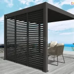 Antracytowa wolnostojąca pergola lamelowa Marlo o kwadratowej podstawie, usytuowana na tarasie z widokiem na morze. Konstrukcja wyposażona jest w pełną zabudowę jednego boku, składającą się z trzech pionowych paneli żaluzjowych z nieruchomymi, poziomymi lamelami. Dach pergoli tworzą ruchome pióra aluminiowe, które na zdjęciu są całkowicie zamknięte. Przy lewym słupie nośnym zamontowany jest mechanizm korbowy służący do ręcznej regulacji zadaszenia. Pod pergolą, na podłożu z jasnoszarych desek, znajdują się dwa nowoczesne fotele z ażurowymi korpusami i jasnymi poduszkami. Taras ograniczony jest białą barierką z wypełnieniem szklanym, za którą widoczna jest linia brzegowa oraz błękitna woda pod pogodnym niebem. Po lewej stronie na horyzoncie miasto położone na wzgórzu, a po prawej błękitne morze.