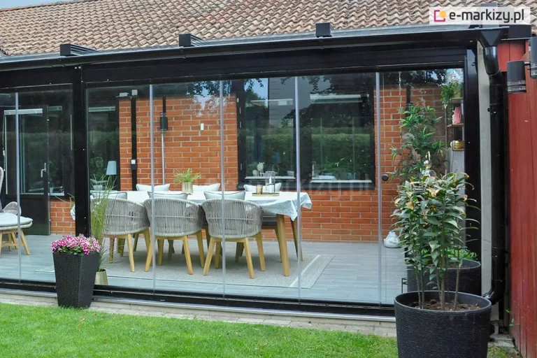 Przeszklona jadalnia tarasowa z systemem bezramowych, przesuwnych szyb Miros, osadzonych w czarnej aluminiowej konstrukcji, przylegająca do budynku z elewacją z czerwonej cegły. Wewnątrz znajduje się drewniany stół nakryty białym obrusem, otoczony sześcioma krzesłami z jasnoszarym siedziskiem i oparciem z plecionki. Na stole ustawiono ozdobne doniczki z roślinami. Podłogę wyłożono jasną deską kompozytową, a całość uzupełnia dywan w części centralnej. Na zewnątrz widoczny zadbany trawnik oraz duże, czarne donice z roślinnością i kwiatami.