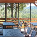 Panoramiczna zabudowa tarasu restauracyjnego wykonana w systemie ścian przesuwnych model Sotro, która integruje wnętrze lokalu z otaczającą naturą. Szklane panele ramowe osadzone są w subtelnych, ciemnych profilach aluminiowych, tworząc transparentną barierę chroniącą przed warunkami atmosferycznymi bez ograniczania widoku na zalesiony krajobraz górski. Konstrukcja wspiera się na drewnianym zadaszeniu z widocznymi belkami, a wewnątrz przestrzeni ustawione są nowoczesne meble ogrodowe, w tym ciemne stoły z listew i dopasowane krzesła z oparciami. Przesuwne skrzydła szklane zachodzą na siebie, co pozwala na elastyczne otwieranie i zamykanie strefy gastronomicznej w zależności od pogody. Całość instalacji podkreśla nowoczesny design obiektu, łącząc funkcjonalność szklanej zabudowy z naturalną estetyką drewnianej balustrady i tarasu, tworząc komfortowe miejsce dla gości restauracji.