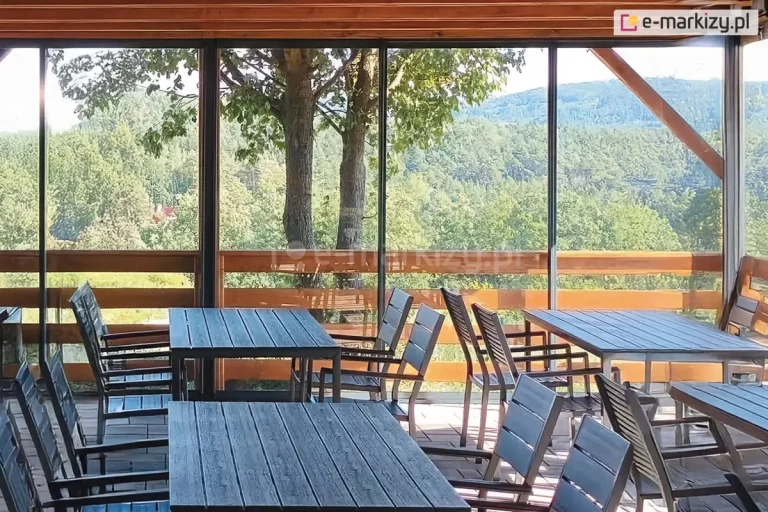 Panoramiczna zabudowa tarasu restauracyjnego wykonana w systemie ścian przesuwnych model Sotro, która integruje wnętrze lokalu z otaczającą naturą. Szklane panele ramowe osadzone są w subtelnych, ciemnych profilach aluminiowych, tworząc transparentną barierę chroniącą przed warunkami atmosferycznymi bez ograniczania widoku na zalesiony krajobraz górski. Konstrukcja wspiera się na drewnianym zadaszeniu z widocznymi belkami, a wewnątrz przestrzeni ustawione są nowoczesne meble ogrodowe, w tym ciemne stoły z listew i dopasowane krzesła z oparciami. Przesuwne skrzydła szklane zachodzą na siebie, co pozwala na elastyczne otwieranie i zamykanie strefy gastronomicznej w zależności od pogody. Całość instalacji podkreśla nowoczesny design obiektu, łącząc funkcjonalność szklanej zabudowy z naturalną estetyką drewnianej balustrady i tarasu, tworząc komfortowe miejsce dla gości restauracji.