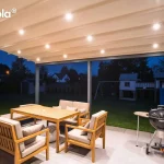 Widok na nowoczesną pergolę Elegancy Tarasola w nocy, z wyraźnie wyeksponowanym systemem oświetlenia LED zintegrowanym z poszyciem dachowym. Jasna tkanina zadaszenia jest w pełni rozwinięta, a liczne punkty świetlne o ciepłej barwie równomiernie oświetlają całą przestrzeń tarasu. Pod zadaszeniem znajduje się komplet drewnianych mebli ogrodowych z jasnymi poduszkami oraz czarny grill węglowy. Konstrukcja pergoli w kolorze antracytowym opiera się na dwóch solidnych profilach przednich, a brak ścian bocznych otwiera widok na ciemny ogród, w którym w tle majaczy podświetlony domek zabaw dla dzieci ze zjeżdżalnią. Całość tworzy nastrojową, wieczorną strefę relaksu przy domu jednorodzinnym.