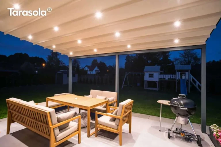 Widok na nowoczesną pergolę Elegancy Tarasola w nocy, z wyraźnie wyeksponowanym systemem oświetlenia LED zintegrowanym z poszyciem dachowym. Jasna tkanina zadaszenia jest w pełni rozwinięta, a liczne punkty świetlne o ciepłej barwie równomiernie oświetlają całą przestrzeń tarasu. Pod zadaszeniem znajduje się komplet drewnianych mebli ogrodowych z jasnymi poduszkami oraz czarny grill węglowy. Konstrukcja pergoli w kolorze antracytowym opiera się na dwóch solidnych profilach przednich, a brak ścian bocznych otwiera widok na ciemny ogród, w którym w tle majaczy podświetlony domek zabaw dla dzieci ze zjeżdżalnią. Całość tworzy nastrojową, wieczorną strefę relaksu przy domu jednorodzinnym.
