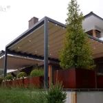 Zdjęcie prezentuje luksusową, nowoczesną pergolę tarasową Tarasola Flat o konstrukcji aluminiowej w kolorze antracytowym, zamontowaną do elewacji piętrowego budynku. Zadaszenie stanowi jasny, kremowy dach tkaninowy zintegrowany z systemem oświetlenia LED w postaci punktowych oczek świetlnych rozmieszczonych na profilach poprzecznych. Konstrukcja wspiera się na wysokich, masywnych słupach i osłania przestronny taras, na którym ustawiono duże, geometryczne donice z cortenu w kolorze rdzawym, wypełnione starannie przyciętymi krzewami oraz trawami ozdobnymi. Budynek posiada nowoczesną architekturę z białym tynkiem, elementami drewnianej stolarki okiennej oraz szklanymi balustradami na wyższej kondygnacji. W tle widoczna jest dalsza część tarasu z kolejnym modułem zadaszenia, co podkreśla modułowy charakter systemu i możliwość osłonięcia bardzo dużych powierzchni wypoczynkowych. Całość aranżacji podświetlona jest ciepłym światłem, tworząc elegancką atmosferę w pochmurny dzień.