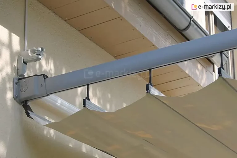 Zdjęcie prezentuje szczegół techniczny montażu ściennego pergoli dachowej Silva na jasnej, tynkowanej elewacji budynku. Głównym elementem jest aluminiowy uchwyt mocujący w kolorze srebrno-szarym, przykręcony bezpośrednio do ściany za pomocą widocznych śrub montażowych. Od uchwytu odchodzi podłużna prowadnica boczna o profilu prostokątnym, która podtrzymuje system zadaszenia. Pod prowadnicą podwieszona jest tkanina osłonowa w kolorze beżowym, zamocowana do poziomych profili przeciwwietrznych za pomocą czarnych uchwytów ślizgowych. Materiał układa się w charakterystyczne fale. Powyżej punktu montażu widoczna jest podbitka dachowa wykonana z jasnobrązowych paneli drewnopodobnych oraz fragment rynny deszczowej. Nad głównym mocowaniem znajduje się również dodatkowy, mniejszy element konstrukcyjny z tworzywa lub metalu, będący częścią systemu sterowania lub stabilizacji. Całość ukazuje solidne połączenie konstrukcji nośnej z bryłą obiektu.