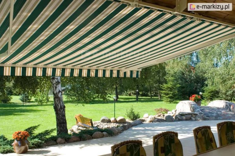 Szeroka markiza Australia Selt w klasycznej, pasiastej kolorystyce, zamontowana nad rozległym tarasem wypoczynkowym w ogrodzie. Poszycie markizy wykonane jest z wytrzymałej tkaniny w regularne, pionowe pasy w kolorach ciemnej zieleni, beżu oraz bieli, co nadaje osłonie tradycyjny i elegancki wygląd. Materiał jest wykończony prostą falbaną, która powtarza wzór pasów, dodatkowo chroniąc przed niskim słońcem. Konstrukcja wsporcza markizy, w tym ramiona przegubowe i rura nawojowa, wykonana jest z aluminium lakierowanego na kolor kremowy, idealnie komponujący się z jasną podłogą tarasu. Markiza Australia Selt jest w pełni rozłożona, rzucając głęboki cień na strefę jadalnianą, w której widoczne są oparcia stylowych krzeseł z technorattanu z kwiecistymi poduszkami. Taras graniczy bezpośrednio z bogato urządzonym ogrodem, w którym znajduje się zadbany trawnik, wysoka brzoza o charakterystycznej białej korze oraz skalniaki z polnych kamieni. W tle widoczna jest dalsza część ogrodu z iglakami, ozdobnymi krzewami oraz małym kamiennym mostkiem. Całość kompozycji oświetla naturalne, letnie słońce, podkreślając funkcjonalność markizy w tworzeniu komfortowej przestrzeni do relaksu na świeżym powietrzu.