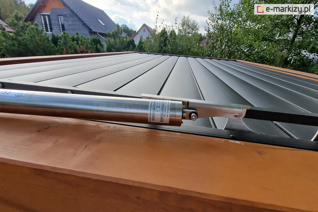 Techniczne zbliżenie na siłownik liniowy sterujący pracą dachu lamelowego w modelu Selt SB400. Srebrny, metalowy napęd o cylindrycznym kształcie jest zamontowany poziomo na krawędzi konstrukcji i połączony z mechanizmem obrotowym lameli za pomocą metalowego trzpienia i śruby. Pod napędem znajduje się fragment ramy wykonanej z drewna o naturalnym, jasnobrązowym odcieniu. Same lamele dachu są w kolorze antracytowym i znajdują się w pozycji całkowicie zamkniętej, tworząc szczelną i płaską powierzchnię zadaszenia. W tle, powyżej linii dachu, widoczne są korony drzew liściastych oraz fragmenty domów jednorodzinnych o skośnych dachach, rozmieszczone na pagórkowatym terenie pod lekko zachmurzonym niebem.