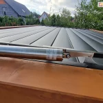 Techniczne zbliżenie na siłownik liniowy sterujący pracą dachu lamelowego w modelu Selt SB400. Srebrny, metalowy napęd o cylindrycznym kształcie jest zamontowany poziomo na krawędzi konstrukcji i połączony z mechanizmem obrotowym lameli za pomocą metalowego trzpienia i śruby. Pod napędem znajduje się fragment ramy wykonanej z drewna o naturalnym, jasnobrązowym odcieniu. Same lamele dachu są w kolorze antracytowym i znajdują się w pozycji całkowicie zamkniętej, tworząc szczelną i płaską powierzchnię zadaszenia. W tle, powyżej linii dachu, widoczne są korony drzew liściastych oraz fragmenty domów jednorodzinnych o skośnych dachach, rozmieszczone na pagórkowatym terenie pod lekko zachmurzonym niebem.