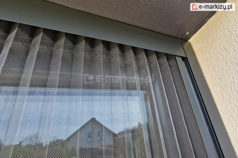 Zbliżenie szarej siatki moskitiery plisowanej Top Zag wsuniętej w profil górny i boczny w kolorze antracytowym zamocowane do wnęki okiennej o jasnej elewacji i kasety rolet zewnętrznych.