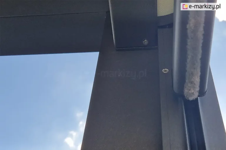 Zbliżenie na detal konstrukcyjny prowadnicy pionowej refleksola Ziiip 120 Selt. Zdjęcie wykonane z perspektywy „od dołu” na tle błękitnego nieba z lekkimi chmurami. Na pierwszym planie widoczna jest czarna, matowa listwa dolna aluminiowa o profilu ceowym, wewnątrz której zamontowana jest gęsta szczotka przeciwkurzowa (uszczelka szczotkowa) w kolorze jasnoszarym. Szczotka ta zapewnia dodatkową izolację i wyciszenie pracy systemu. W górnej części kadru widoczny jest fragment połączenia prowadnicy z kasetą rolety oraz śruba montażowa z łbem wpuszczanym. Konstrukcja charakteryzuje się precyzyjnym wykończeniem i nowoczesną, surową estetyką w kolorze antracytowym.