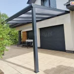 Zdjęcie prezentuje perspektywiczny widok na nowoczesne zadaszenie tarasowe Guma, obecnie Deponti, w kolorze antracytowym, skupiając się na przednim słupie nośnym i konstrukcji dachu. Jednospadowy dach wypełniony jest półprzezroczystymi panelami z poliwęglanu, które są zamocowane pomiędzy aluminiowymi krokwiami. Konstrukcja posiada zintegrowany system rynnowy, którego zakończenie na szczycie słupa zostało zabezpieczone estetyczną, zaokrągloną maskownicą z logo producenta. W tle widoczny jest taras wyłożony beżowymi płytami, jasna elewacja domu z zamkniętymi czarnymi roletami zewnętrznymi oraz fragmenty zielonych krzewów ogrodowych.