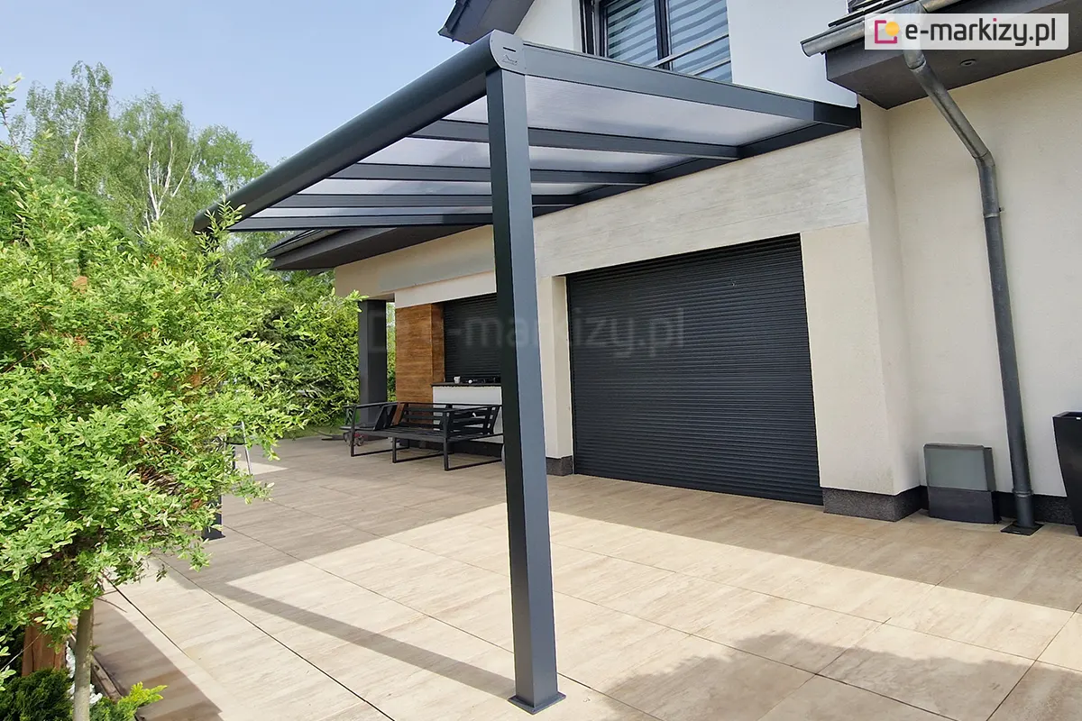 Zdjęcie prezentuje perspektywiczny widok na nowoczesne zadaszenie tarasowe Guma, obecnie Deponti, w kolorze antracytowym, skupiając się na przednim słupie nośnym i konstrukcji dachu. Jednospadowy dach wypełniony jest półprzezroczystymi panelami z poliwęglanu, które są zamocowane pomiędzy aluminiowymi krokwiami. Konstrukcja posiada zintegrowany system rynnowy, którego zakończenie na szczycie słupa zostało zabezpieczone estetyczną, zaokrągloną maskownicą z logo producenta. W tle widoczny jest taras wyłożony beżowymi płytami, jasna elewacja domu z zamkniętymi czarnymi roletami zewnętrznymi oraz fragmenty zielonych krzewów ogrodowych.