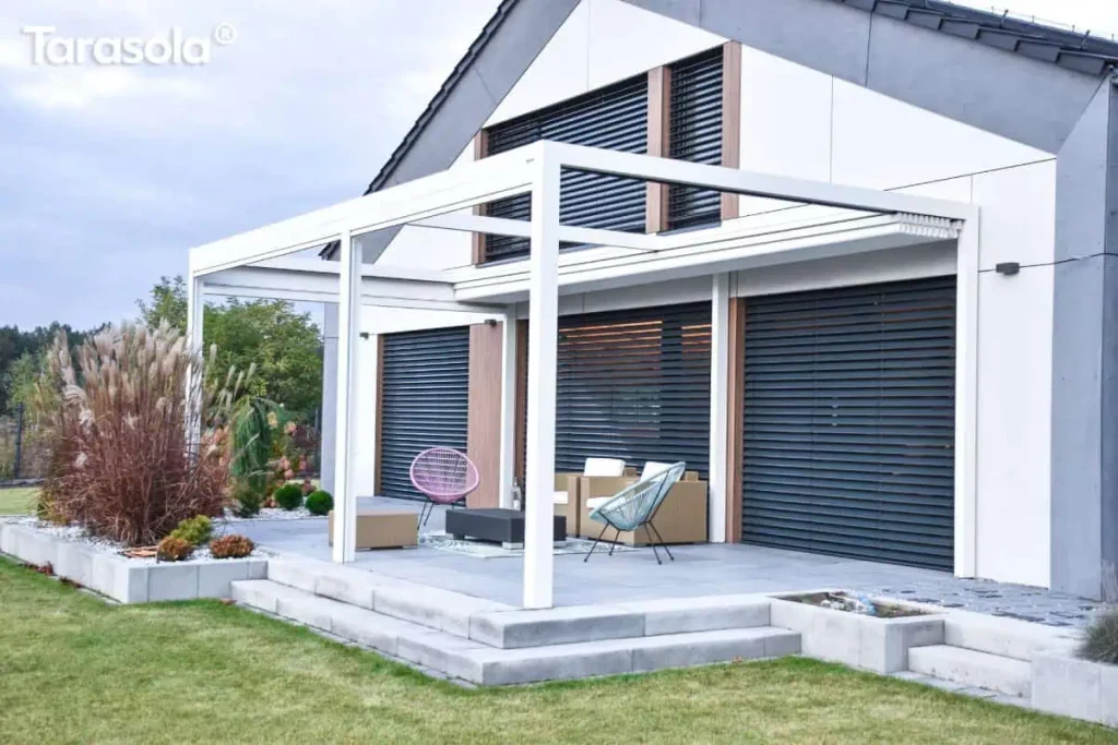 Zdjęcie przedstawia wolnostojącą pergolę tarasową Tarasola Flat o aluminiowej konstrukcji lakierowanej na kolor śnieżnobiały, zamontowaną na nowoczesnym tarasie z szarych płyt betonowych. Zadaszenie tkaninowe pergoli jest w tym momencie całkowicie zwinięte, co odsłania minimalistyczną ramę i poprzeczne profile prowadzące. Taras jest wielopoziomowy, wykończony stopniami z jasnoszarych bloczków i otoczony pasem dekoracyjnego białego grysiku z nasadzeniami traw ozdobnych oraz niskich krzewów. Na tarasie zaaranżowano strefę wypoczynkową z nowoczesnymi meblami, w tym beżowymi fotelami o kubikowym kształcie oraz designerskimi krzesłami typu Acapulco w kolorze różowym i błękitnym, ustawionymi wokół niskiego, czarnego stolika kawowego. Budynek w tle ma białą elewację z szarymi wstawkami oraz duże przeszklenia osłonięte zewnętrznymi żaluzjami fasadowymi w kolorze grafitowym, które są całkowicie zamknięte. Perspektywa zdjęcia z poziomu ogrodu podkreśla kontrast między jasną konstrukcją pergoli a ciemnymi elementami wykończenia fasady domu.