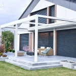 Zdjęcie przedstawia wolnostojącą pergolę tarasową Tarasola Flat o aluminiowej konstrukcji lakierowanej na kolor śnieżnobiały, zamontowaną na nowoczesnym tarasie z szarych płyt betonowych. Zadaszenie tkaninowe pergoli jest w tym momencie całkowicie zwinięte, co odsłania minimalistyczną ramę i poprzeczne profile prowadzące. Taras jest wielopoziomowy, wykończony stopniami z jasnoszarych bloczków i otoczony pasem dekoracyjnego białego grysiku z nasadzeniami traw ozdobnych oraz niskich krzewów. Na tarasie zaaranżowano strefę wypoczynkową z nowoczesnymi meblami, w tym beżowymi fotelami o kubikowym kształcie oraz designerskimi krzesłami typu Acapulco w kolorze różowym i błękitnym, ustawionymi wokół niskiego, czarnego stolika kawowego. Budynek w tle ma białą elewację z szarymi wstawkami oraz duże przeszklenia osłonięte zewnętrznymi żaluzjami fasadowymi w kolorze grafitowym, które są całkowicie zamknięte. Perspektywa zdjęcia z poziomu ogrodu podkreśla kontrast między jasną konstrukcją pergoli a ciemnymi elementami wykończenia fasady domu.