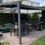 Zdjęcie przedstawia dużą, wolnostojącą pergolę tarasową Square o nowoczesnej konstrukcji z profili aluminiowych w kolorze ciemnoantracytowym, zlokalizowaną w przydomowym ogrodzie. Zadaszenie pergoli wykonane jest z jasnego, wodoodpornego materiału, który został całkowicie rozsunięty, odsłaniając poprzeczne belki wzmacniające. Pod pergolą, na podłożu wyłożonym wielobarwną kostką brukową w odcieniach szarości i brązu, zaaranżowano rozbudowaną strefę relaksu. W centralnej części znajduje się stół jadalniany z ciemnego technorattanu ze szklanym blatem, otoczony czterema krzesłami z tego samego materiału. W głębi ustawiono duży narożnik ogrodowy z ciemnobrązowej plecionki, wyposażony w liczne, grube poduszki oparciowe w pasującym kolorze. Obok narożnika na bruku leży duży pies o ciemnej sierści. Po lewej stronie widoczna jest zielona huśtawka ogrodowa z miękkim siedziskiem oraz fragment nowoczesnej wanny spa (jacuzzi) w obudowie z ciemnego drewna lub kompozytu, na której brzegu stoją kieliszki oraz butelka szampana. W tle znajduje się wysoki, gęsty żywopłot oddzielający posesję od sąsiedniego, jasnego budynku mieszkalnego, a po prawej stronie widoczne są pnącza na pergoli bocznej oraz zadbany trawnik. Konstrukcja pergoli opiera się na kilku solidnych pionowych słupach, a całość tworzy komfortową, zadaszoną przestrzeń do wypoczynku na świeżym powietrzu.