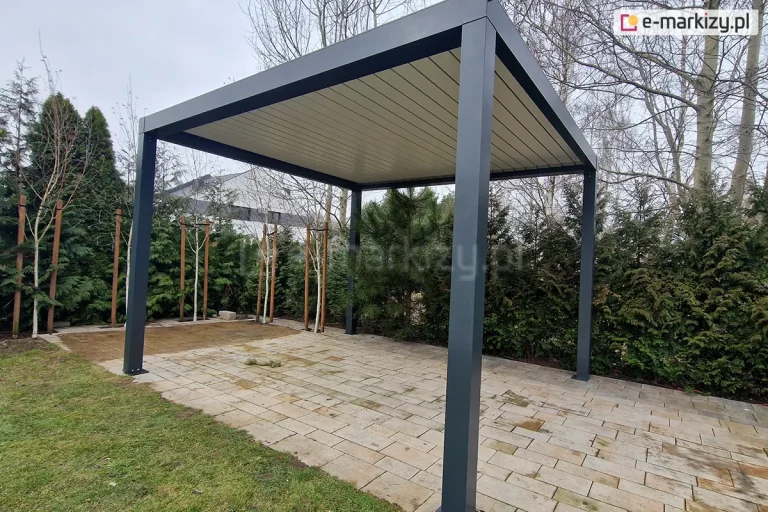 Wolnostojąca pergola aluminiowa SB350 w kolorze antracytowym, zamontowana na podłożu z jasnej, prostokątnej kostki brukowej. Konstrukcja posiada cztery solidne słupy nośne o kwadratowym przekroju oraz płaski dach z ruchomymi lamelami w kontrastującym, białym kolorze, które są obecnie szczelnie zamknięte. W tle widoczny jest ogród z gęstym żywopłotem z iglaków, młodymi drzewkami liściastymi wspartymi na palikach oraz fragmentem elewacji nowoczesnego budynku mieszkalnego. Zdjęcie wykonane z niskiej perspektywy, ukazujące detale mocowania stóp pergoli do podłoża oraz wykończenie górnej ramy.
