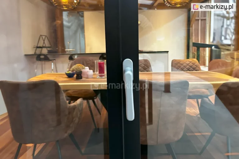 Zbliżenie na środkowy zamek klamkowy zamontowany na pionowym profilu aluminiowym bezramowego systemu przesuwnego Auris. Zdjęcie prezentuje minimalistyczną klamkę o ergonomicznym kształcie, osadzoną na antracytowym profilu domykającym tafle szkła. Przez przezroczyste przeszklenie widoczne jest stylowe wnętrze jadalni z dużym drewnianym stołem, nowoczesnymi krzesłami tapicerowanymi oraz dekoracjami stołowymi. W tle widać fragment kuchni oraz ciepłe oświetlenie wiszących lamp. Detal podkreśla estetykę wykończenia okuć oraz funkcjonalność bezpiecznego zamykania dużych powierzchni przeszklonych.