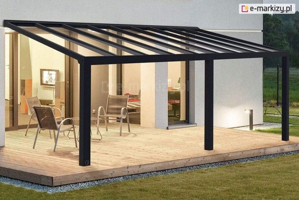 Nowoczesne, stałe zadaszenie tarasowe Modes Glas w kolorze czarnym lub głębokiego antracytu, zamontowane do białej elewacji budynku. Konstrukcja dachu opiera się na gęstym układzie równoległych, aluminiowych krokwi, które podtrzymują transparentne tafle szkła bezpiecznego, hartowanego. Całość wsparta jest na dwóch solidnych, kwadratowych słupach nośnych usytuowanych na krawędzi tarasu. Przestrzeń pod zadaszeniem została wyłożona szerokimi, drewnianymi deskami o naturalnym odcieniu, na których ustawiono trzy metalowe krzesła ażurowe ze srebrzystym wykończeniem oraz niewielki stolik kawowy. Wewnątrz domu, widocznym przez duże przeszklenia, dostrzec można ciepłe oświetlenie salonu, a na zewnątrz, przy krawędzi tarasu, znajduje się niska lampa ogrodowa oświetlająca zielony trawnik o zmierzchu.