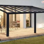 Nowoczesne, stałe zadaszenie tarasowe Modes Glas w kolorze czarnym lub głębokiego antracytu, zamontowane do białej elewacji budynku. Konstrukcja dachu opiera się na gęstym układzie równoległych, aluminiowych krokwi, które podtrzymują transparentne tafle szkła bezpiecznego, hartowanego. Całość wsparta jest na dwóch solidnych, kwadratowych słupach nośnych usytuowanych na krawędzi tarasu. Przestrzeń pod zadaszeniem została wyłożona szerokimi, drewnianymi deskami o naturalnym odcieniu, na których ustawiono trzy metalowe krzesła ażurowe ze srebrzystym wykończeniem oraz niewielki stolik kawowy. Wewnątrz domu, widocznym przez duże przeszklenia, dostrzec można ciepłe oświetlenie salonu, a na zewnątrz, przy krawędzi tarasu, znajduje się niska lampa ogrodowa oświetlająca zielony trawnik o zmierzchu.