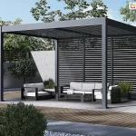 Nowoczesna wolnostojąca pergola ogrodowa Marlo Duo w kolorze antracytowym, której kluczowym elementem są zintegrowane panele boczne w formie poziomych żaluzji. Ta aluminiowa konstrukcja z dachem lamelowym została wyposażona w stałe przesłony boczne z wypełnieniem panelami żaluzjowymi, które harmonizują z grafitowym stelażem, zapewniając skuteczną ochronę przed słońcem, wiatrem oraz zwiększając prywatność użytkowników. Pod zadaszeniem i przed żaluzjową ścianką znajduje się minimalistyczny zestaw mebli tarasowych z jasnymi poduszkami, ustawiony na podłożu z płyt betonowych i drewna. Całość aranżacji dopełnia betonowy murek oraz zadbana roślinność ogrodowa, podkreślająca techniczny i elegancki design paneli bocznych.