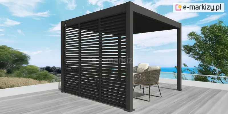 Grafika przedstawia wizualizację nowoczesnej, wolnostojącej pergoli tarasowej o konstrukcji aluminiowej w kolorze ciemnego antracytu, umieszczonej na przestronnym tarasie z widokiem na morze. Głównym elementem dekoracyjnym i funkcjonalnym widocznym na pierwszym planie jest system trzech paneli bocznych wypełnionych poziomymi lamelami. Panele te, utrzymane w tej samej kolorystyce co rama konstrukcyjna, tworzą ażurową ścianę, która pozwala na regulację dopływu światła oraz zapewnia ochronę przed wiatrem i wzrokiem sąsiadów. Pod zadaszeniem pergoli zaaranżowano strefę wypoczynkową, w której znajdują się dwa stylowe fotele o plecionej strukturze z jasnymi, miękkimi poduszkami na siedziskach i oparciach. Meble ustawione są na podłodze tarasu wykonanej z szerokich desek kompozytowych w kolorze jasnego, chłodnego drewna. W tle rozpościera się malowniczy krajobraz z linią brzegową, błękitną wodą oraz zieloną roślinnością nadmorską pod jasnym, lekko zachmurzonym niebem. Taras zabezpieczony jest nowoczesną balustradą ze szklanym wypełnieniem, co podkreśla luksusowy i otwarty charakter prezentowanej architektury ogrodowej.