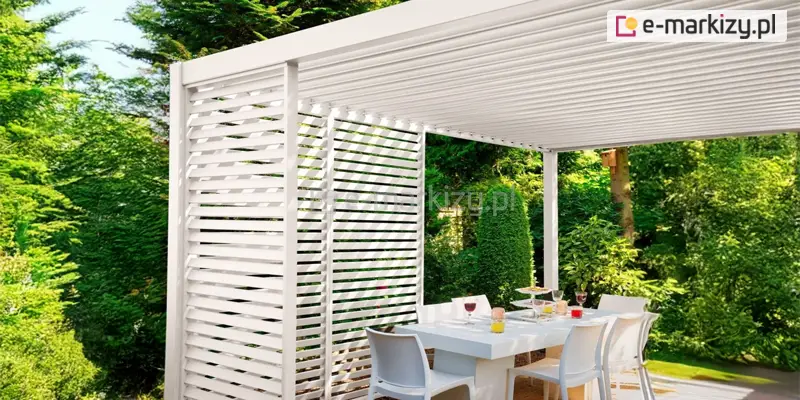Zdjęcie przedstawia nowoczesną pergolę ogrodową w kolorze białym, pod którą zaaranżowano jadalnię na świeżym powietrzu. Konstrukcja posiada płaski dach wykonany z ruchomych, poziomych lameli, które na zdjęciu są zamknięte. Dwie ze ścian bocznych pergoli zostały częściowo osłonięte przez system stałych paneli żaluzjowych. Panele te składają się z szerokich, białych listew zamontowanych pod stałym kątem, co tworzy efekt ażurowej ściany zapewniającej cień i prywatność. Pod zadaszeniem znajduje się duży, biały stół o prostej formie, przy którym ustawione są nowoczesne, białe krzesła z tworzywa. Na stole widoczne są nakrycia, kieliszki z winem oraz szklanki z napojami, co sugeruje przygotowanie do posiłku. Pergola jest usytuowana w otoczeniu intensywnej, soczystej zieleni ogrodu – w tle widoczne są gęste krzewy, wysokie drzewa iglaste oraz liściaste, które tworzą naturalną, izolującą ścianę roślinności. Światło słoneczne przenikające przez liście i konstrukcję nadaje całej scenie jasny, letni charakter.
