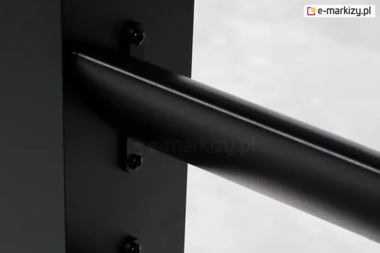 Zbliżenie na element montażowy stałego panelu żaluzjowego Louvre w kolorze czarnym, wykonanego z aluminium o matowym wykończeniu. Zdjęcie przedstawia detal mocowania poziomej, eliptycznej lameli do pionowego profilu bocznego. Lamele są przytwierdzone za pomocą czarnych, metalowych uchwytów w kształcie litery L, które stabilnie trzymają profil za pomocą widocznych śrub z łbem sześciokątnym. Tło zdjęcia jest jasne i nieostre, co pozwala skupić uwagę na technicznych szczegółach łączenia oraz precyzji wykonania konstrukcji. Całość prezentuje solidny i estetyczny system montażu bez widocznych spawów.