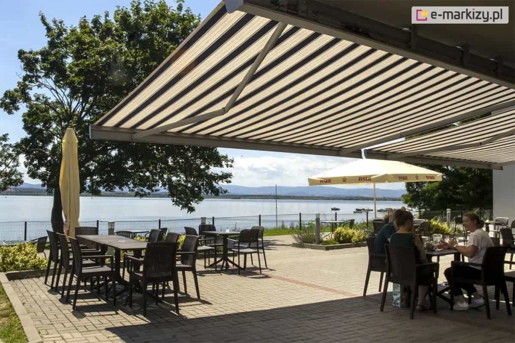 Zdjęcie prezentuje szeroki, zewnętrzny taras restauracyjny położony bezpośrednio nad brzegiem jeziora, w pełni osłonięty przez dwie połączone markizy Corsica. Poszycie markiz posiada klasyczny wzór w szerokie, pionowe pasy w odcieniach beżu i ciemnego brązu, co harmonizuje z naturalnym otoczeniem. Pod zadaszeniem ustawiono liczne stoliki kawowe oraz ciemnobrązowe krzesła z technorattanu. Przy jednym ze stolików siedzą goście, korzystając z cienia w słoneczny dzień. Taras jest wyłożony szarą kostką brukową i oddzielony od jeziora niskim, metalowym ogrodzeniem panelowym. W tle rozpościera się widok na spokojną taflę wody, na której unoszą się małe łodzie, oraz na zarysy gór na horyzoncie pod jasnym niebem. Po lewej stronie tarasu rośnie rozłożyste, zielone drzewo, przy którym stoi złożony parasol ogrodowy w kolorze kremowym. W centralnej części placu widoczny jest również drugi, rozstawiony biały parasol reklamowy. Całość sceny ukazuje funkcjonalne zastosowanie dużych osłon przeciwsłonecznych w branży gastronomicznej, tworzących komfortowe miejsce wypoczynku z widokiem na naturę.